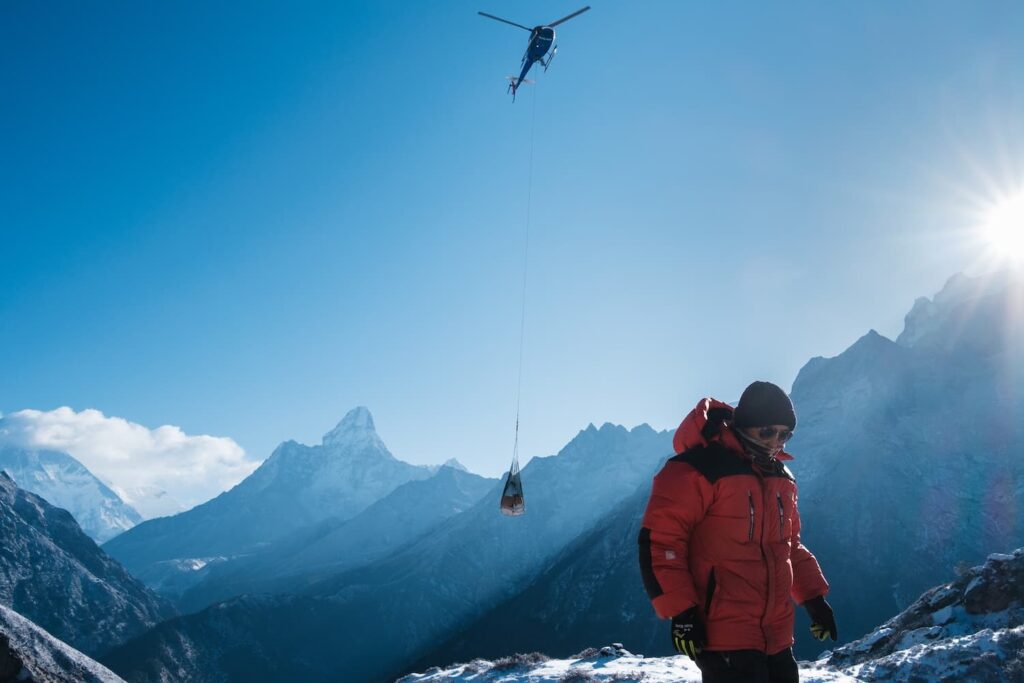 A person in a red jacket stands on a snowy mountain with a helicopter in the sky carrying a load on a cable, surrounded by snow-covered peaks and a bright sun in the background.