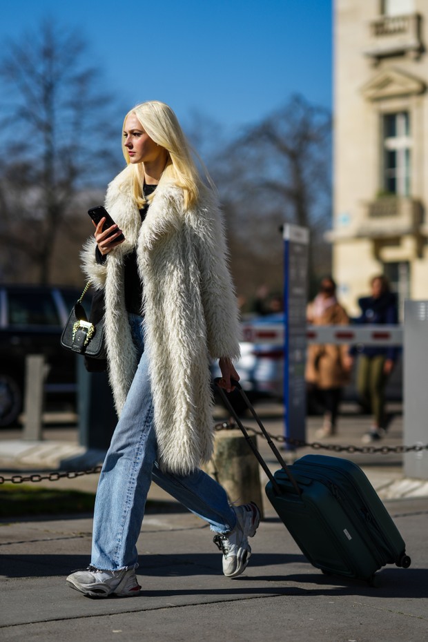 PARIS, FRANCE - MARCH 08: A guest wears a black t-shirt, a white long fluffy coat, blue faded denim wide legs pants, a black shiny leather handbag from Versace, a navy blue suitcase, gray suede and white leather sneakers, outside Chanel, during Paris Fash (Foto: Getty Images)