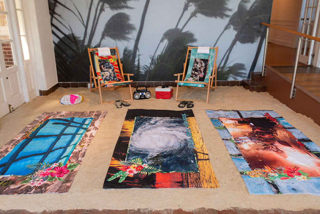 An installation featuring three custom-printed towels on sand in a gallery with two beach chairs.