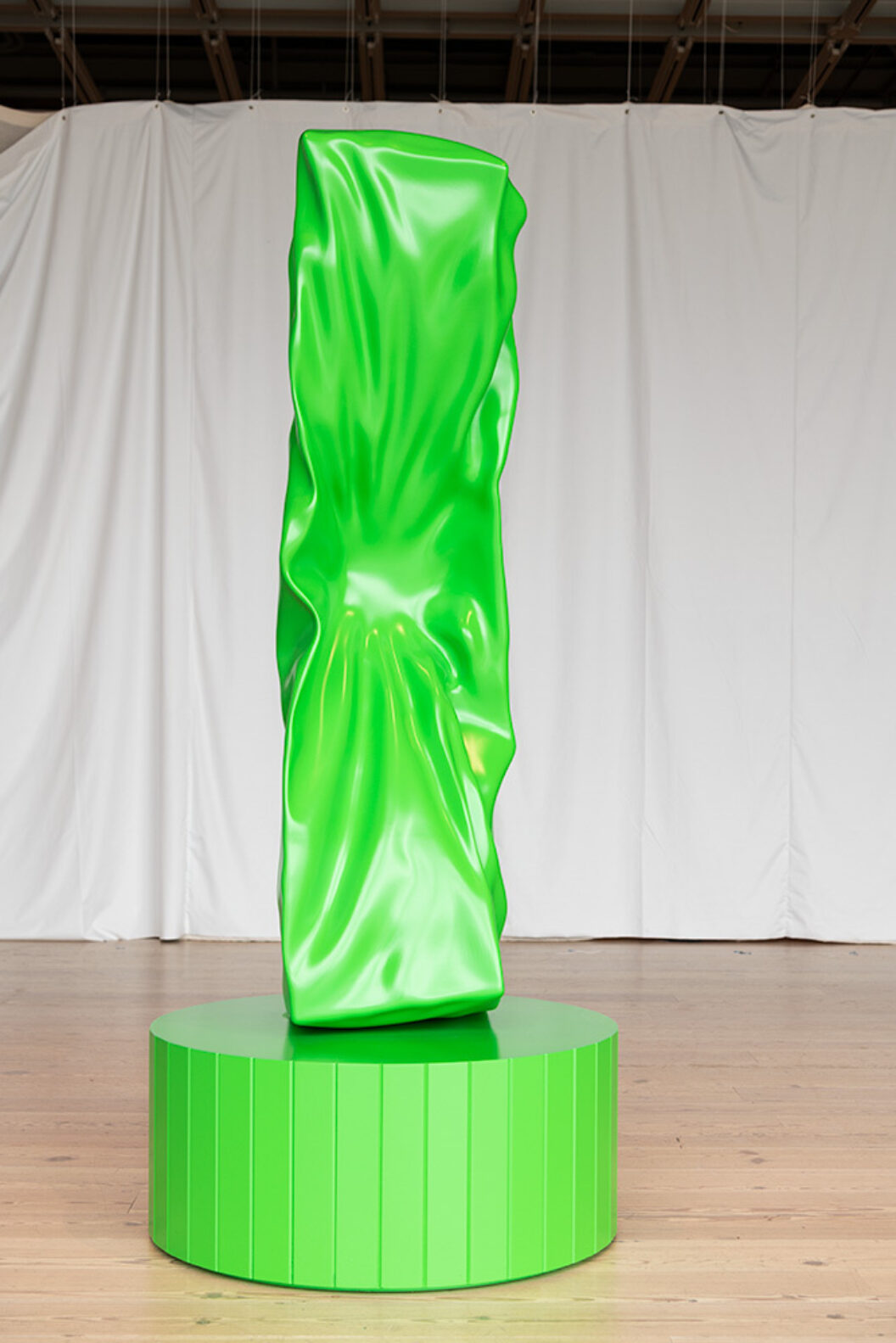 An upright rectangular green sculpture with a compressed dent in its middle photographed against a white curtain.