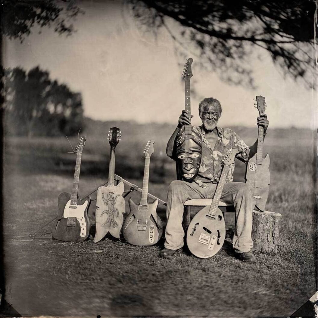 A sepia photograph of an older black man sitting under a tree surrounded by homemade guitars.