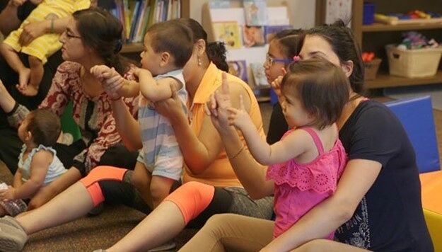 A row of parents sit on the floor with their babies being held up engaged in an activity.