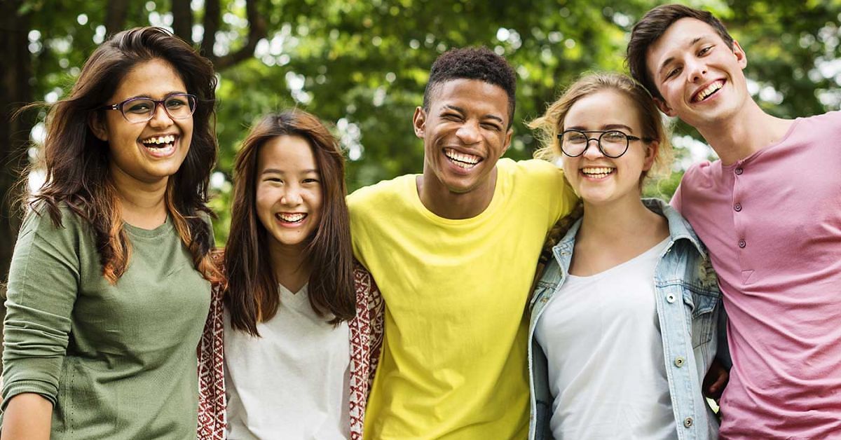 Five teens of a variety of genders and races wearing brightly colored clothes facing the camera and smiling with trees in the background.