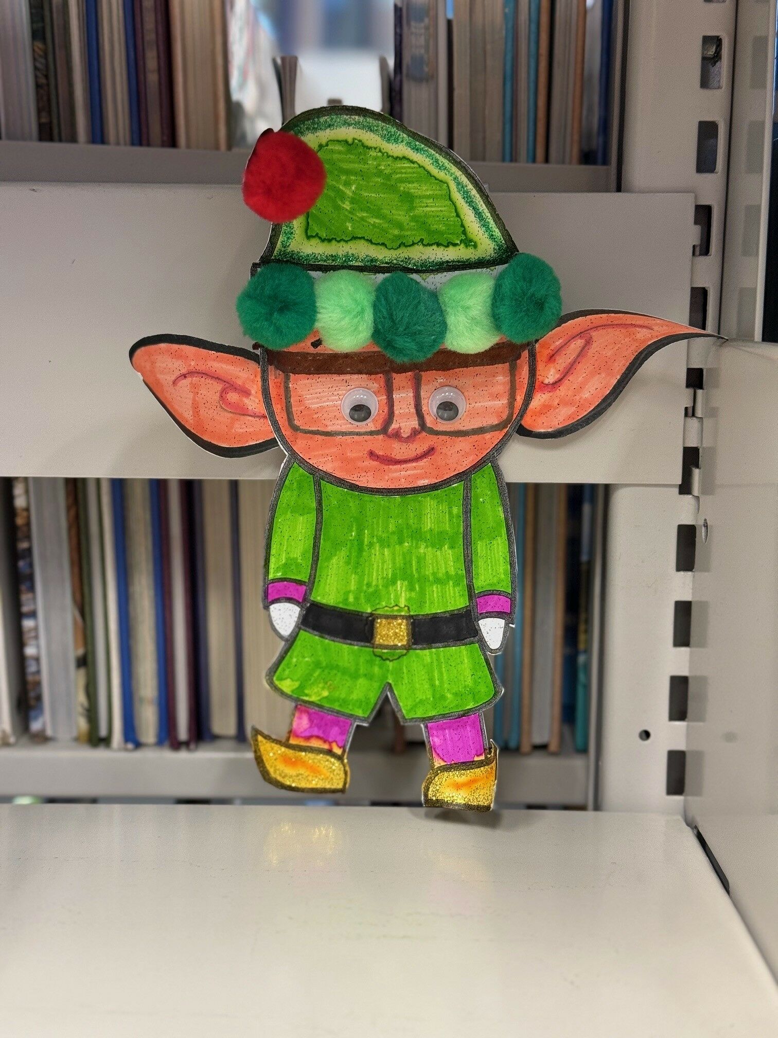 A nerdy elf with glasses and a green outfit hiding on a bookshelf