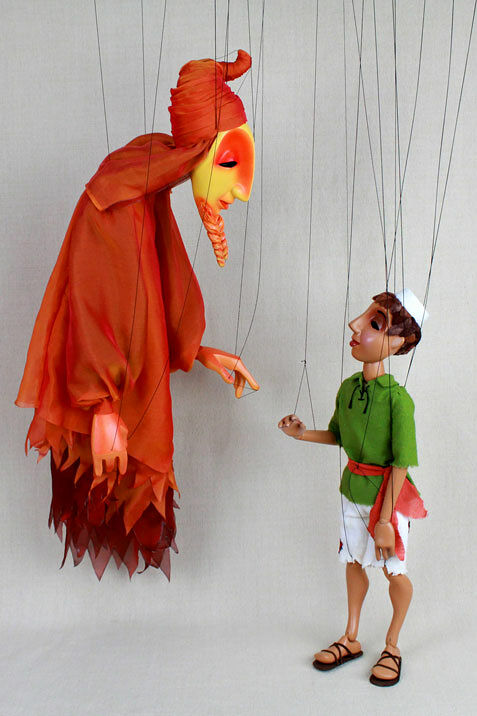 marionette of boy in green shirt, white shorts and dark sandals faving a genie wearing layers of sheer red-orange robes over brown robes