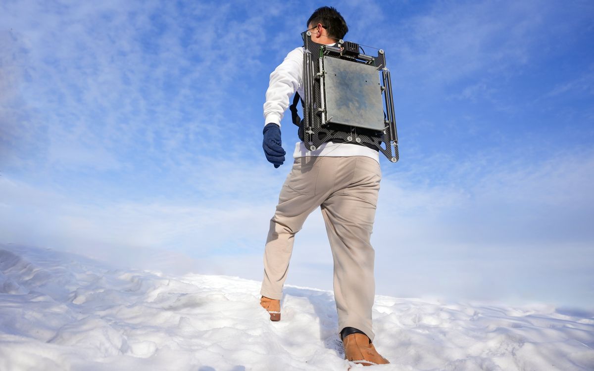 A man walking up a snowy hill wearing a large metal plate with springs and rods over his backpack.