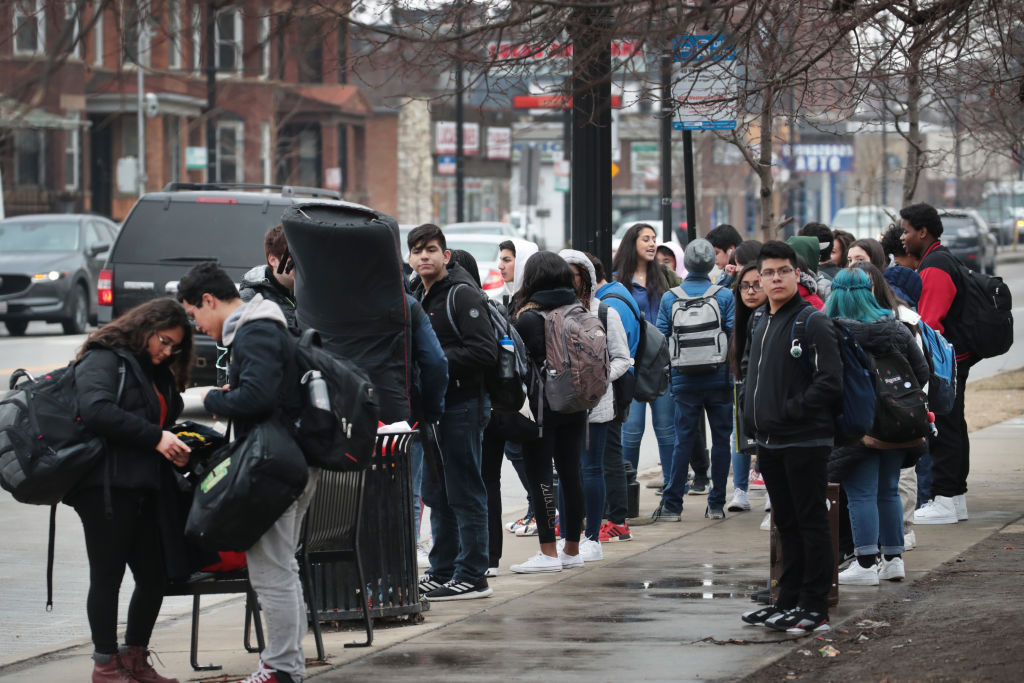 Students stand on a sidewalk waiting for a bus.