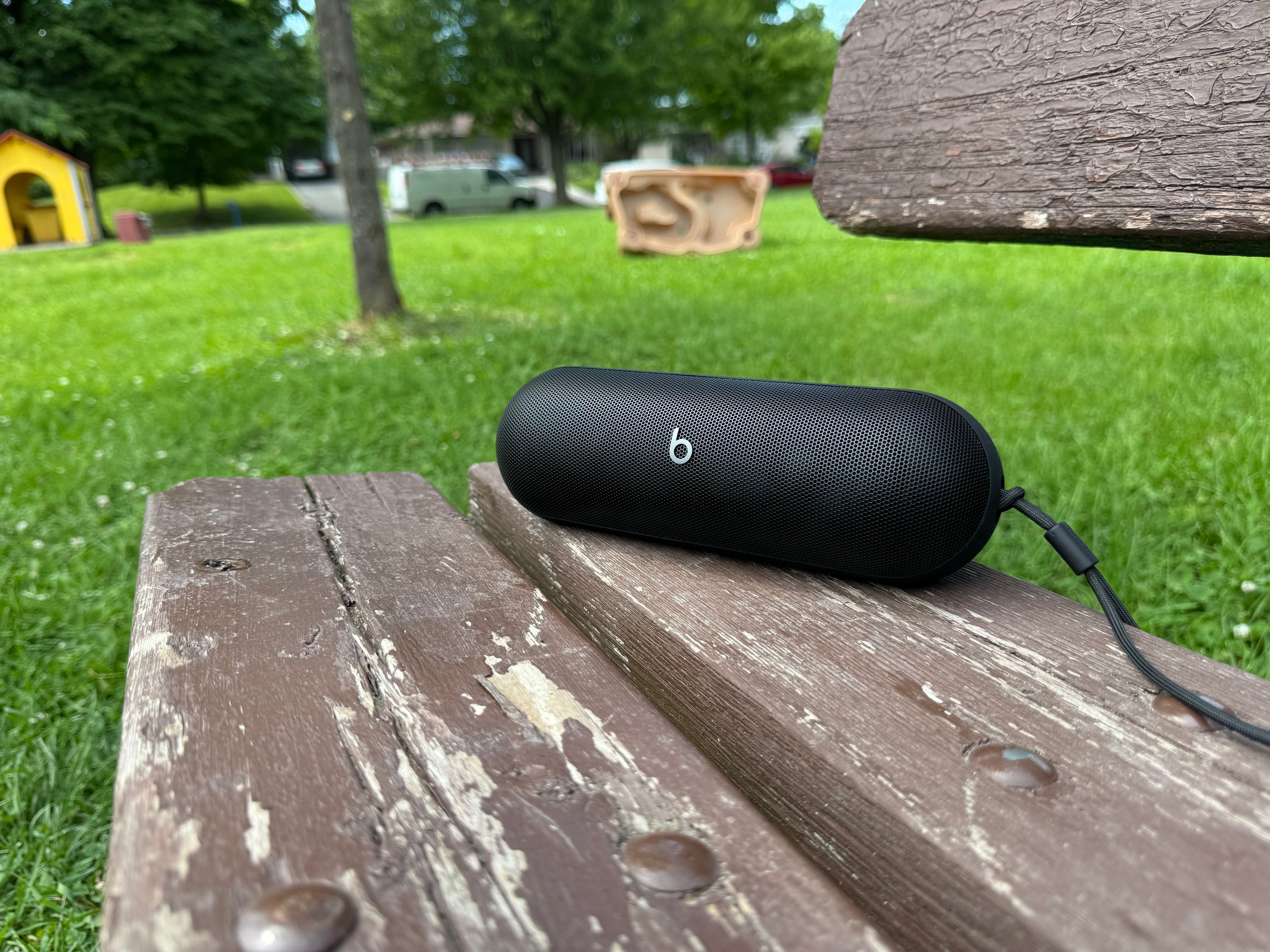 A black 2024 Beats Pill on a brown wooden park bench with green grass and a tree in the background.