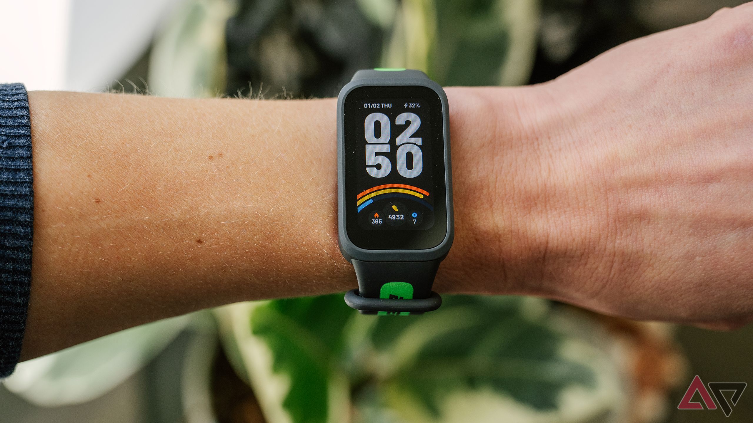 The Xiaomi Smart Band 9 Active is on a wrist held above blurred green plants. 