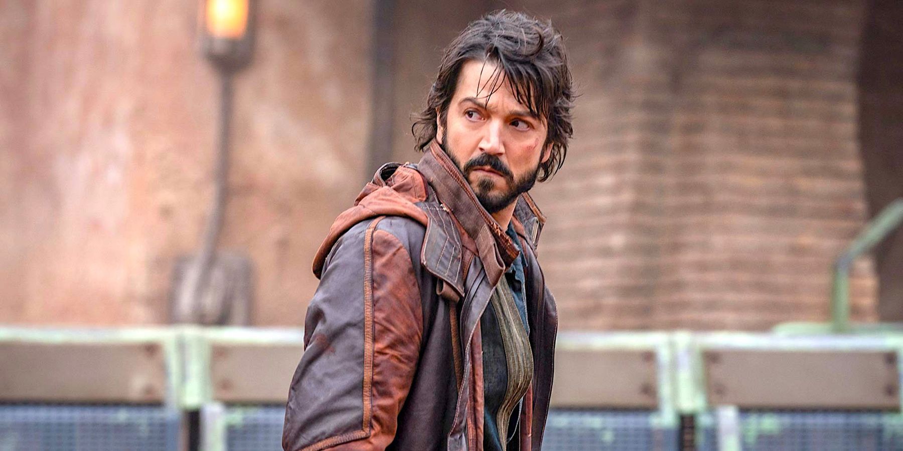 Cassian Andor (Diego Luna), sporting a beard and heavy jacket, walking the streets of Ferrix in Andor.