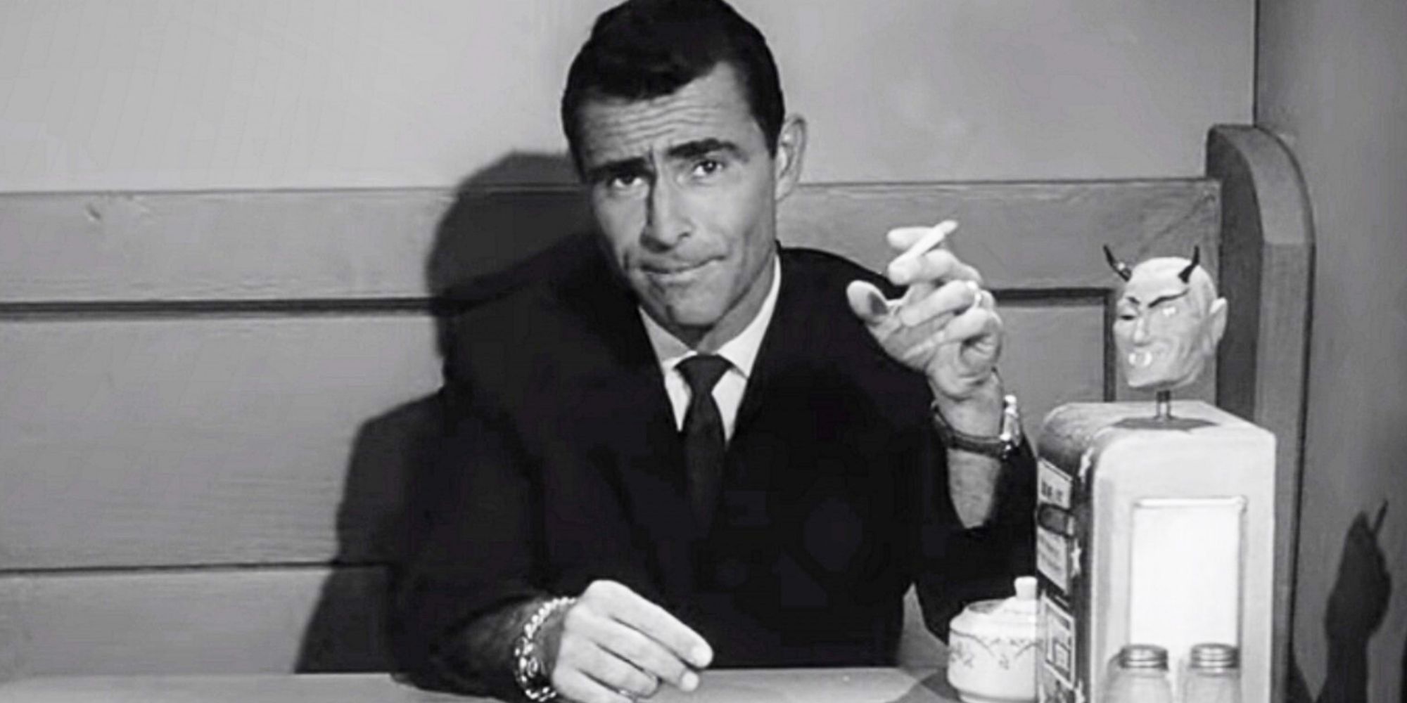 Rod Serling sitting at a table smoking a cigarette in The Twilight Zone.