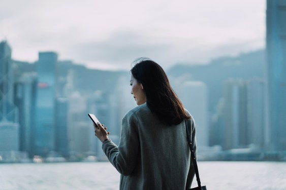 Young Asian businesswoman using smartphone by the promenade of Victoria harbour, against Hong Kong city scene of central business district