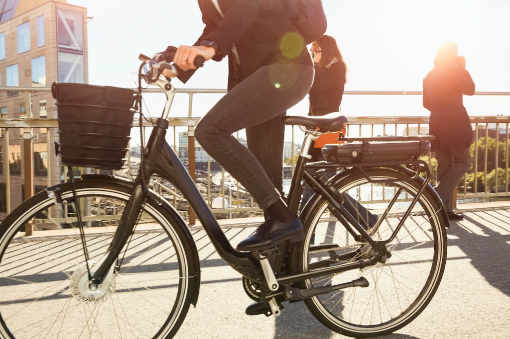 Low section of mature woman riding electric bicycle by commuters on bridge in city against sky