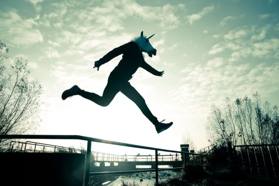 An adult wearing a unicorn mask leaps over a chain-link fence