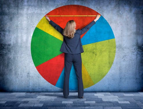 A businesswoman, using a tape measure, reaches up to measure her piece of the pie on a large pie chart that is projected onto a concrete wall.