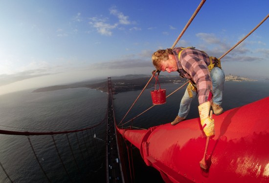 A worker painting the Golden Gate Bridge with San Francisco in the background.