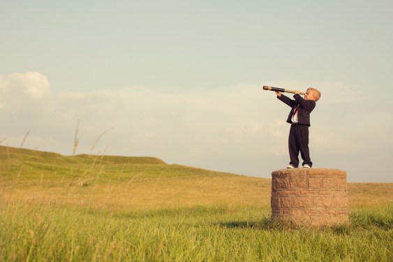 A young British boy in a business suit is standing on a pedestal looking through a hand-held telescope in the English countryside. He is looking for more business opportunities.