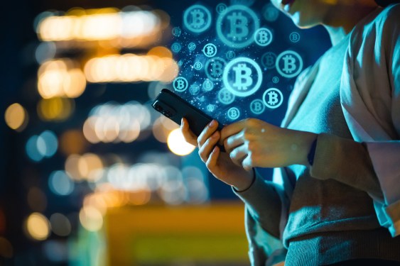 Young East Asian woman shown from the jaw doing using a smartphone, with city lights shown blurred behind and digital illustrations of Bitcoin coming out of the phone