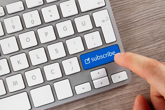 Close-up shot of human finger while it pushes the blue "Subscribe" button on aluminium computer keyboard on office desk.