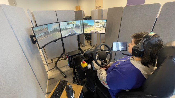 A Vay teleoperator in Las Vegas sits behind a steering wheel viewing 3 large monitors.