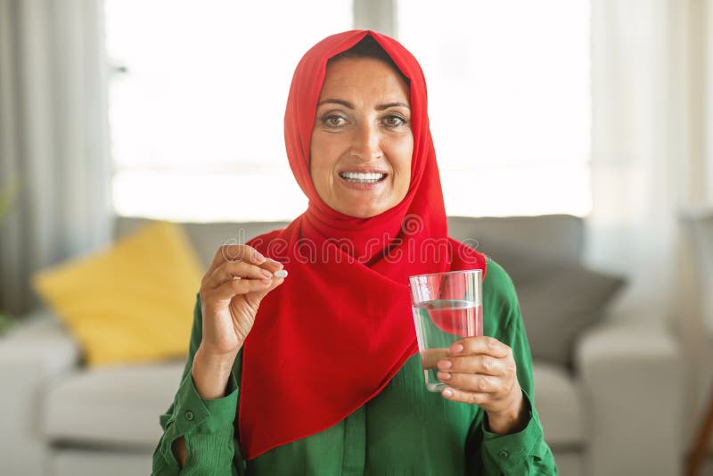 A cheerful woman with her head covered in a vibrant red hijab is holding a pill and a clear glass of water, seemingly preparing to take the medication as she stands inside a well-lit room. A cheerful woman with her head covered in a vibrant red hijab is holding a pill and a clear glass of water, seemingly preparing to take the medication as she stands inside a well-lit room.