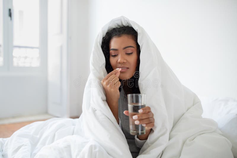 A young Hispanic woman is wrapped snugly in a white comforter, sitting in a sunlit bedroom. She is taking medication, holding a clear glass of water in one hand and a pill in the other. A young Hispanic woman is wrapped snugly in a white comforter, sitting in a sunlit bedroom. She is taking medication, holding a clear glass of water in one hand and a pill in the other