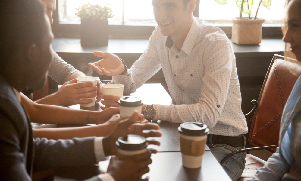 Multi ethnic group of happy friends drinking coffee in paper cups together in cozy cafe, diverse multiracial african and caucasian young people talking sharing table having fun at coffeehouse meeting