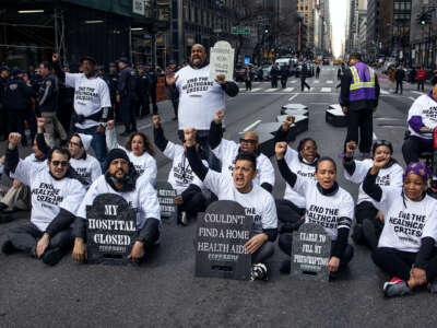 Protesters holding headstone-shaped signs bearing statements like "COULDN'T FIND A HOME HEALTH AID" and "MY HOSPITAL CLOSED" as they block an intersection during an outdoor demonstration