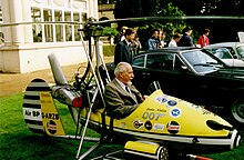 Small, one man, open-cockpit helicopter on a lawn about the size of a car next to it, with a man sitting in it.