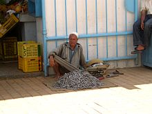 Vendeur de poisson assis sur le trottoir et appuyé contre le mur ; la marchandise est déposée à ses pieds.