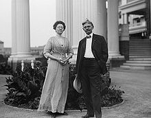 A man posing in front of several large Greek columns. He is about sixty-years-old and is wearing a two piece black suit and bow tie with a hand in his pocket and holding a wide brimmed white hat in the other while standing in a casual pose with a partial smile and his hair being blown up by the wind. A woman stands to his right wearing a full-length gown and holding a folded fan between her clasped hands; her glasses obscure her eyes and while she stands in a dignified pose.