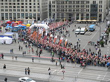 Barevná fotografie s pohledem na průjezd cyklistů obklopených diváky ulicemi městského centra