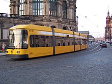 Barevná fotografie s pohledem na projíždějící žlutou tramvaj, za níž je jeden z pavilónů Zwingeru