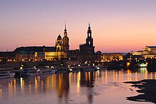 Barevná fotografie s pohledem na osvětlené městské panorama s věžemi kostelů v době setmění