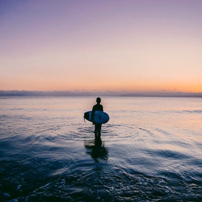 夕暮れの海に佇むサーファーの写真