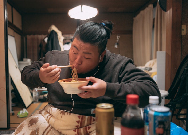 散らかった部屋でラーメンを食べる肥満男子の写真