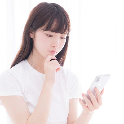 スマホを眺めて考え込む白いTシャツの女性の写真