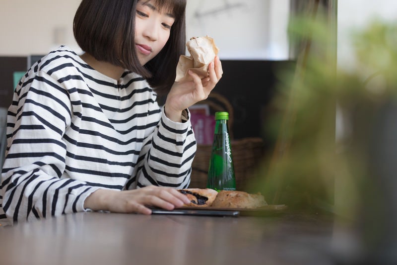 フードコートでパンを食べながらスマホを見る女性の写真