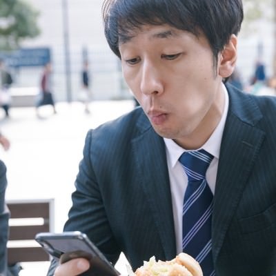 ファーストフードを食べながらスマホをチェックする現代サラリーマンの写真