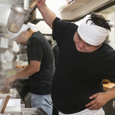 高い位置から湯切りにチャレンジするラーメン店主の写真