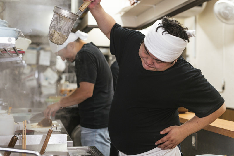 高い位置から湯切りにチャレンジするラーメン店主の写真