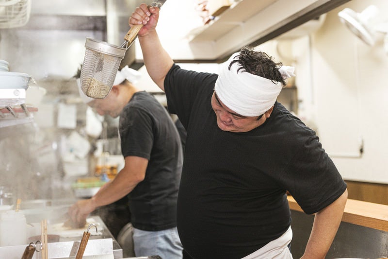 手を上げて勢いよく湯切りするラーメン店主の写真