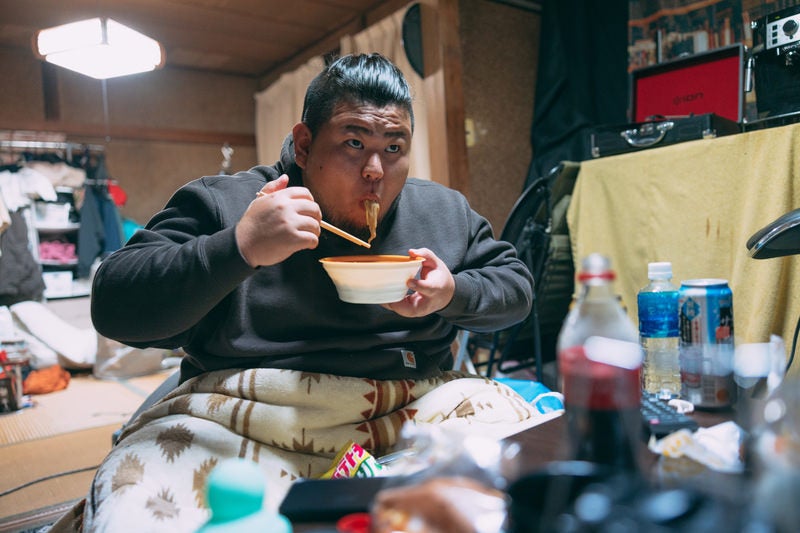 自宅でラーメンを啜る無精髭の男性の写真