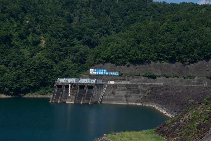 山肌に掲げられた国土交通省とダム名の看板とその下に見える発電所取水口（福井県大野市）の写真