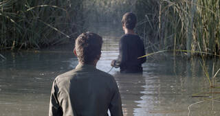 Still from a video showing two boys with their backs to the camera, wading through water towards some reeds. The water is brown and muddy, the sun is shining from the left. 