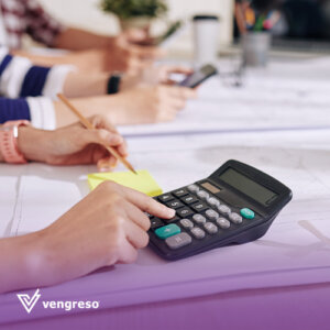 close up of hands typing on a calculator on a desk holding a pencil with other people hands blurred in the background using sales tools