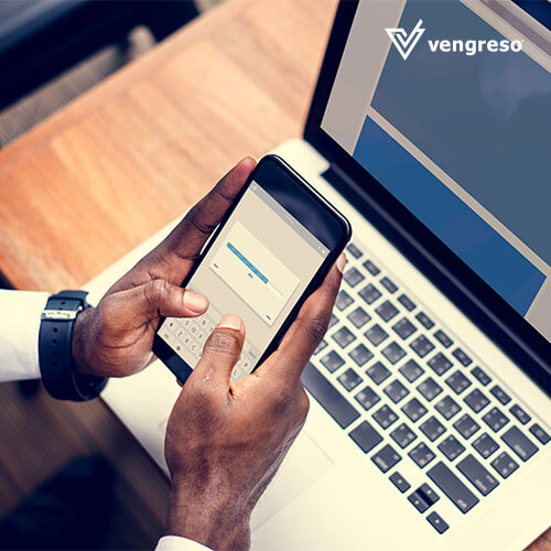 A man is using a cell phone to automate data entry while sitting at a desk with a laptop.