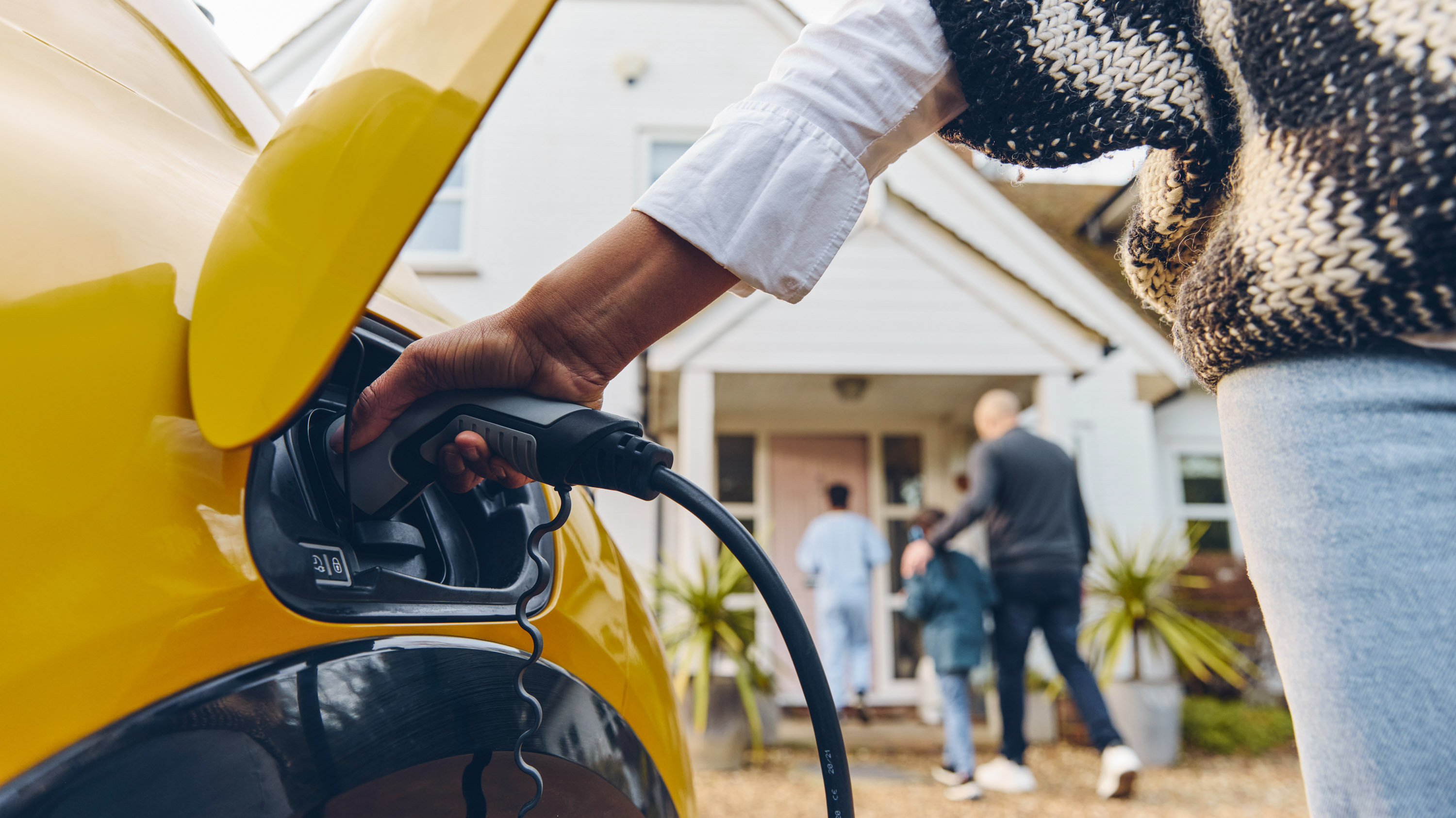 Woman plugging electric charger into car parked in front of a home with three people walking up to the front door