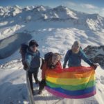 Three people on a snowy mountain top holding a rainbow flag