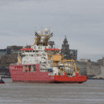 A large red ship on a body of water, with Liverpool city in the background